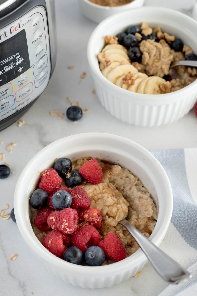 Instant Pot oat meal for one in a white bowl topped with brown sugar, blueberries and raspberries, with another bowl in the background topped with granola, banana, and blueberries, with an Instant Pot in the background.