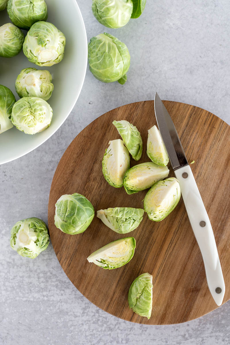 an overhead shot of a small knife cutting pale green brussels sprouts on a round wooden cutting board