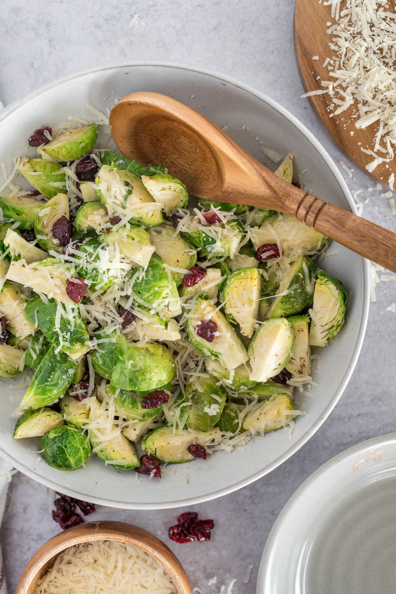 An overhead shot of a bowl of cooked green brussels sprouts topped with parmesan and cranberries and being tossed by a wooden spoon