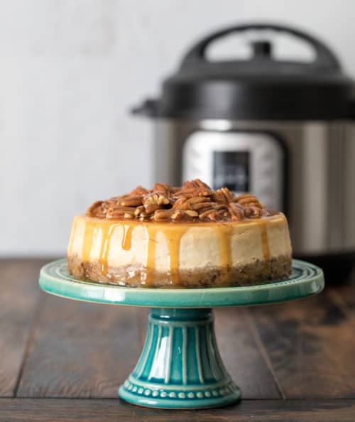 Pressure Cooker / Instant Pot Caramel Pecan Cheesecake on a cake stand in front of an Instant Pot
