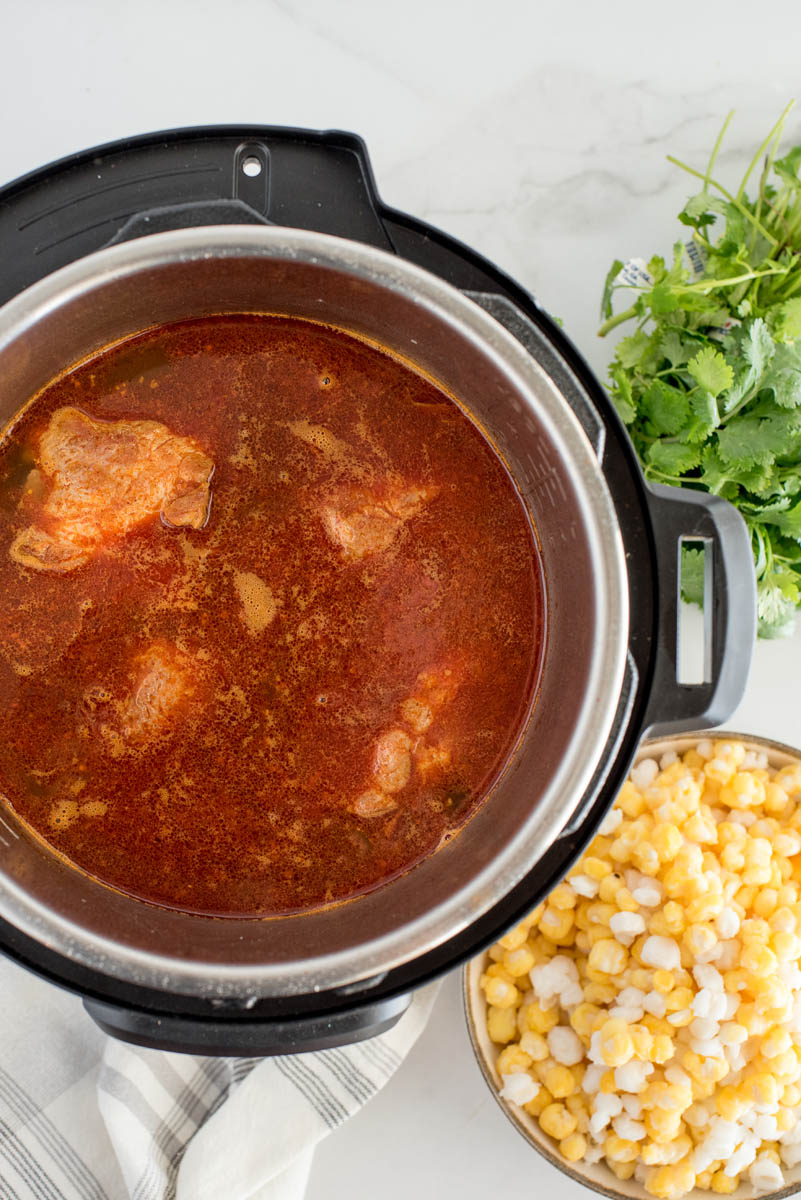 An overhead vertical shot looking into an Instant Pot filled pork and spices and broth to make the stew, with cilantro and hominy alongside the pressure cooker ready to add after cooking
