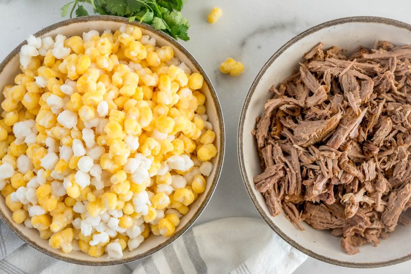 A side-by-side overhead photo of a bowl full of white and yellow hominy and a second bowl filled with shredded pork