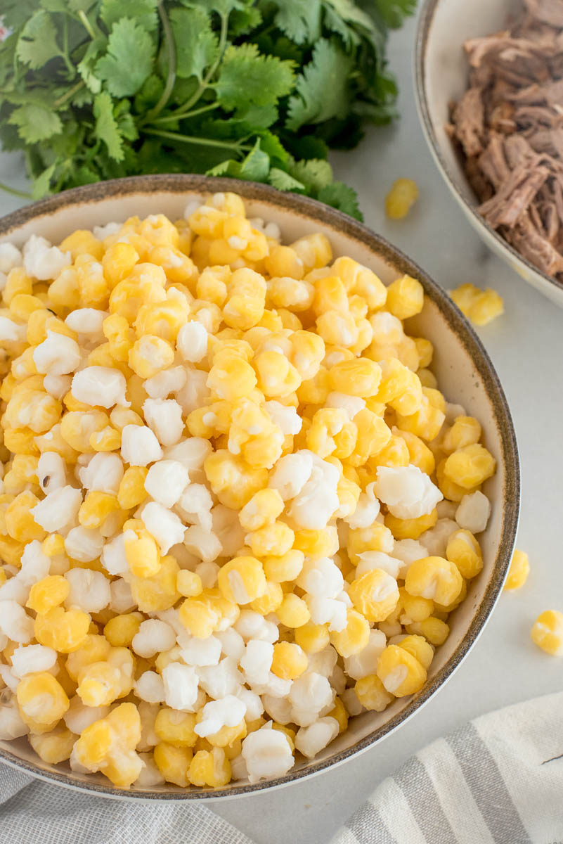 A close-up look at a bowl filled with yellow and white hominy with cilantro and shredded pork in the background