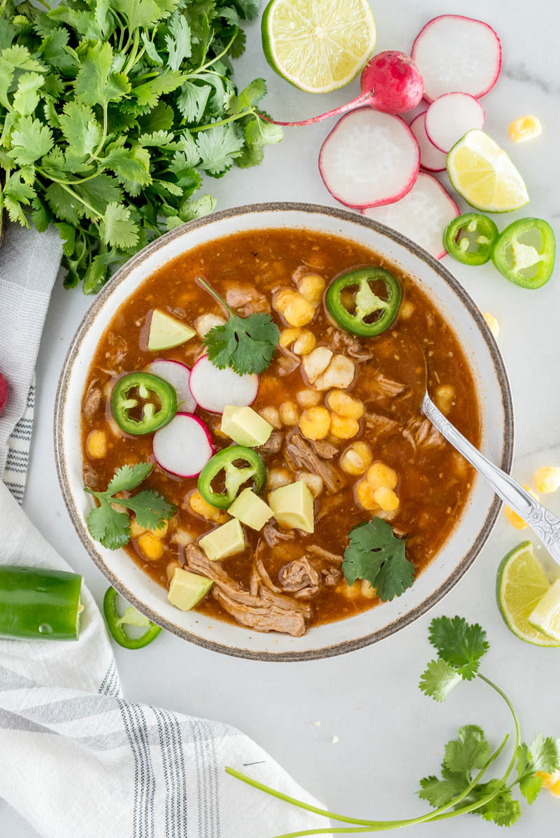 An overhead shot of a white bowl dished up with Instant Pot Pork stew, filled with shredded pork, hominy, radishes, peppers, cilantro, and limes for garnish