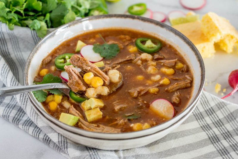a horizontal 45 degree shot of a spoonful of InstaPot Pork Stew with shredded pork, hominy, avocado,, and a slice of cornbread and radishes in the background