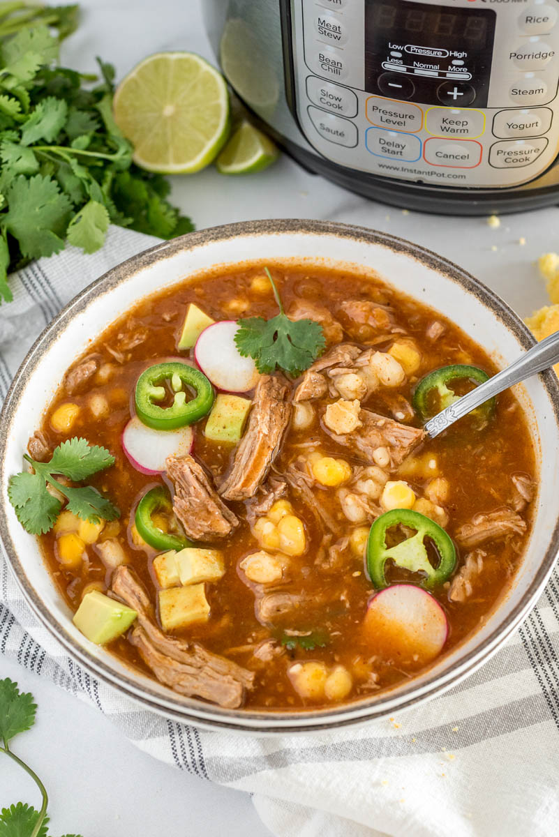 An shot of a white bowl filled with Instant Pot Pork Stew with hominy (quick pozole), with an Instant Pot and cilantro lime garnish in the background