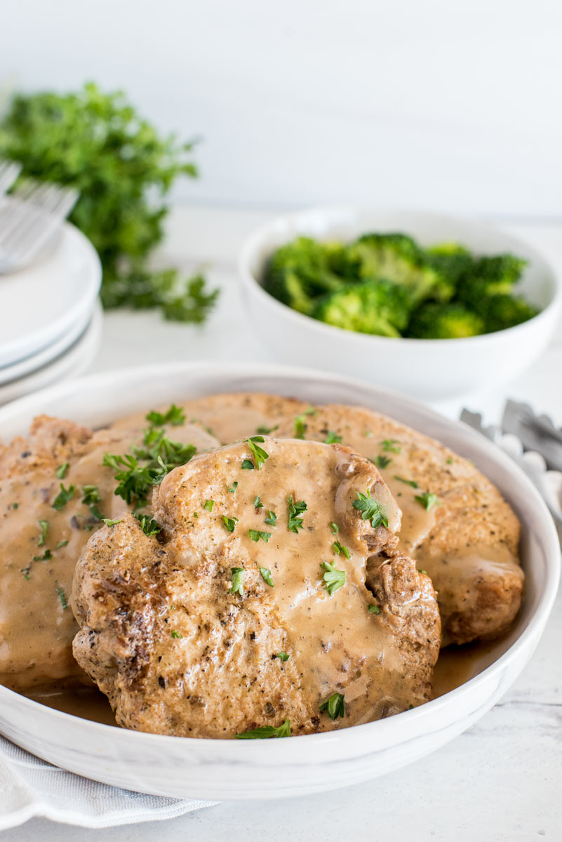 close up on a white bowl with four pork chops in mushroom gravy with steamed broccoli in the background