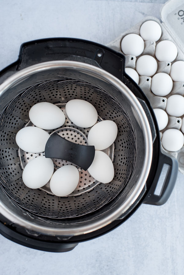 Overhead of An Instant Pot with a steamer basket inside with seven white eggs ready to be hard-boiled with a carton of eggs nearby.