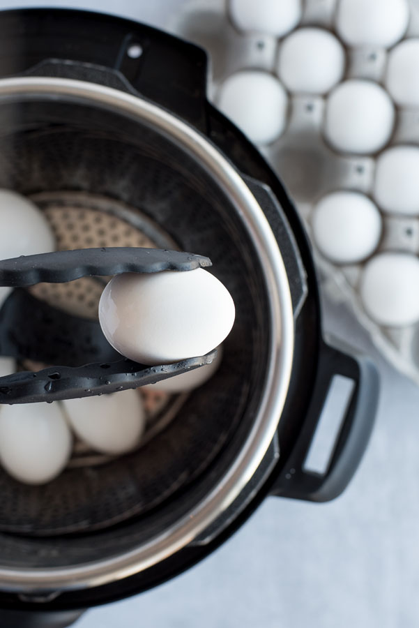 Close up of a white hard-boiled egg coming out of an Instant Pot with black and silver tongs.