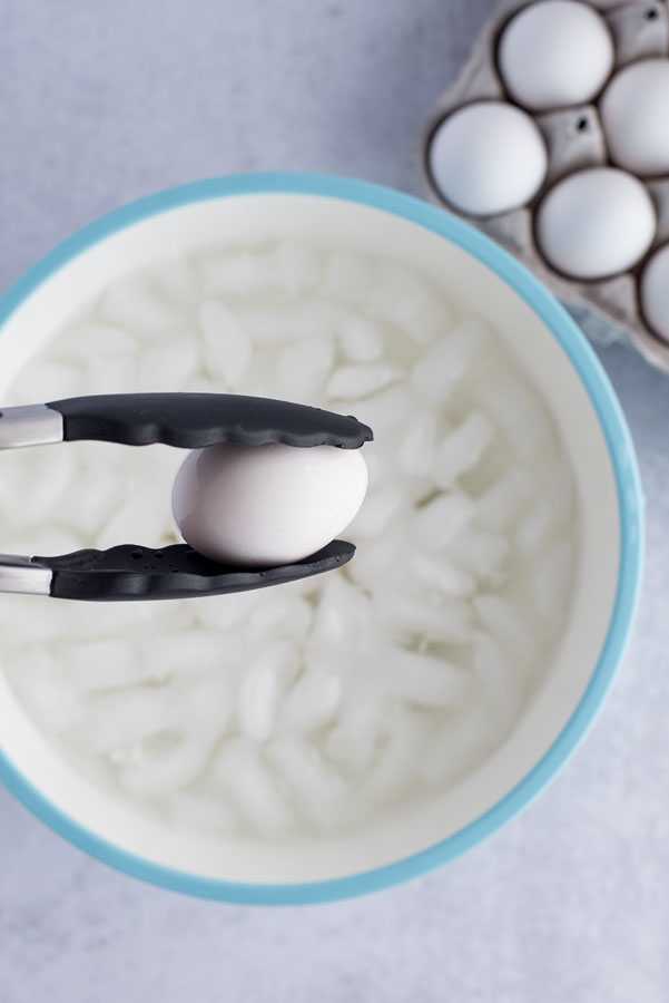 Close up on black and silver tongs holding a white hard-boiled egg over an ice bath after cooking in a pressure cooker / Instant Pot.