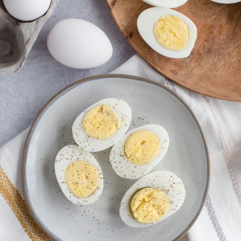 Overhead of a grey ceramic plate with four halves of perfectly cooked Instant Pot / pressure cooker hard-boiled eggs sprinkled with salt and black pepper on a white, grey and yellow striped cloth dishtowel.