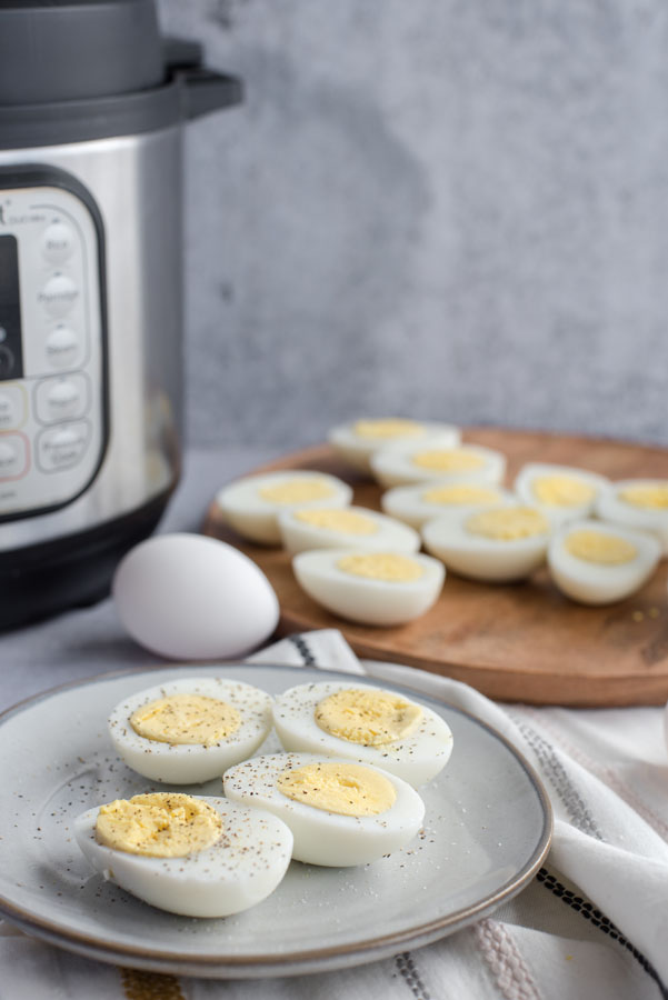 Side view of a grey ceramic plate with four halves of hard-cooked eggs with an Instant Pot and a large wooden tray of eggs in the background.