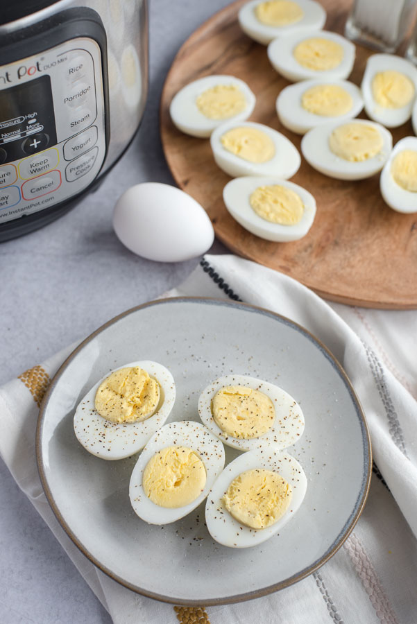 Overhead of a grey ceramic plate with four halves of perfectly cooked hard-boiled eggs with black pepper on a grey slate background with an Instant Pot and tray of more eggs in the background.