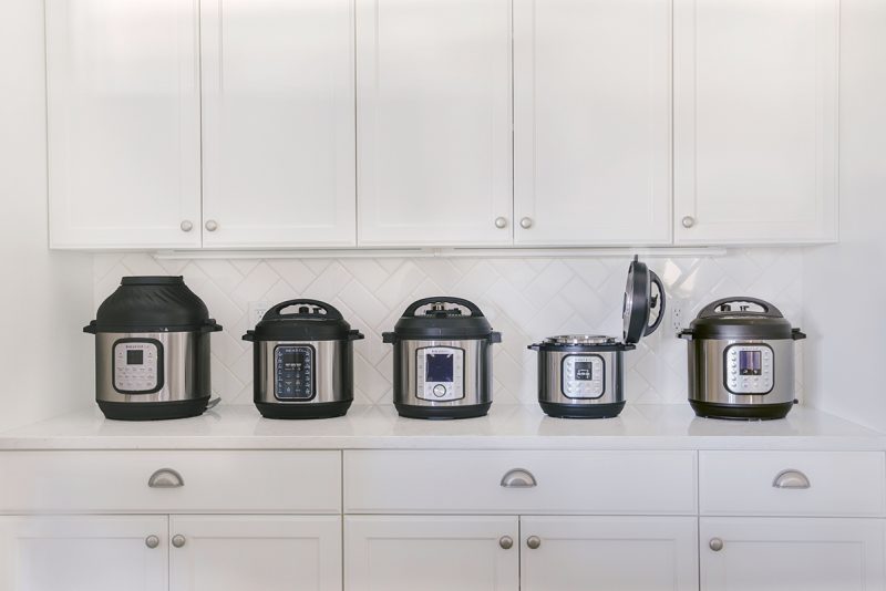 Five different models of Instant Pot on a white counter with white cabinets and a white tile backsplash in the background