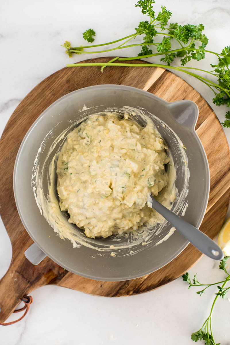 An overhead shot of stirring together the egg salad ingredients, looking into a gray mixing bowl on a wooden round cutting board, with parsley in the background. 
