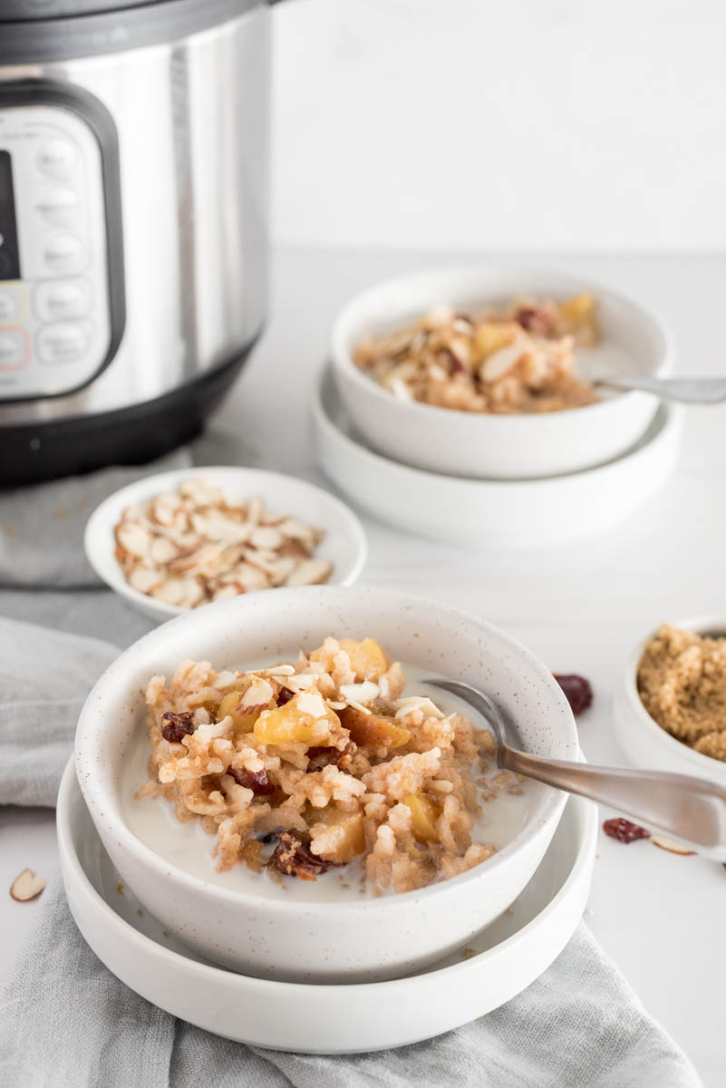 A vertical shot with an Instant Pot and bowls of almonds and cherries in the background with a white speckled bowl filled with breakfast apple risotto, garnished with diced cherries and a splash of milk, with a spoon on the side ready to eat