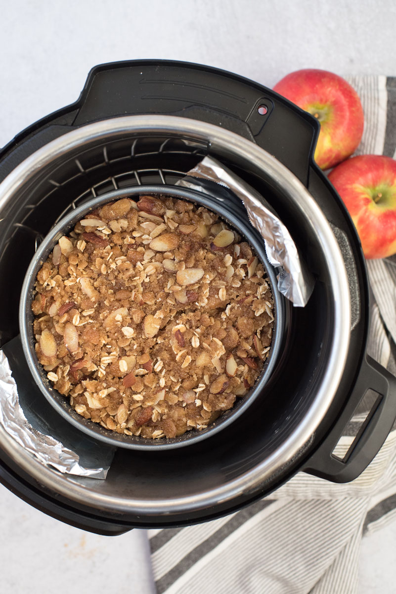 an overhead shot of the instide of the Instant Pot with a foil sling and a springform pan containing finished Instant Pot apple crisp with two red apples in the upper right