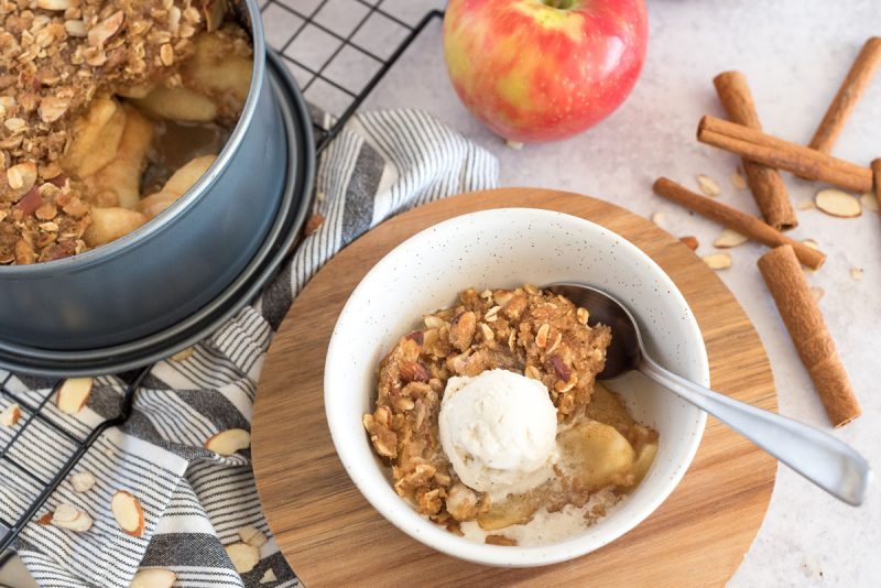 a shot of Instant Pot apple crisp in a white bowl, with a scoop of vanilla ice cream on top, with the pan of apple crisp, apples, and cinnamon sticks in the background