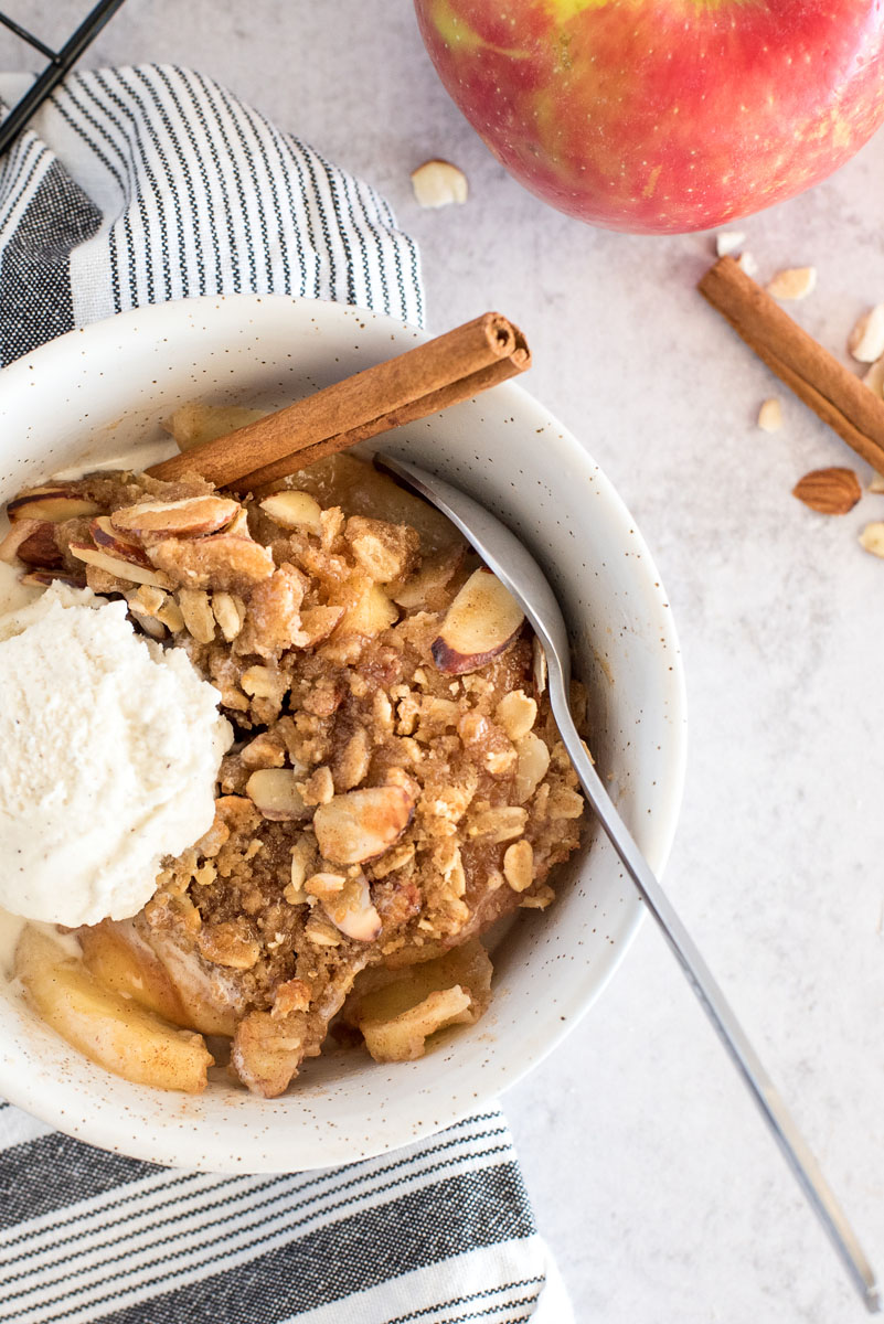 an overhead closeup shot of ninja foodi apple crisp in a white bowl, with a scoop of vanilla ice cream on top, with a silver spoon and a cinnamon stick in the bowl and a red apple in the background