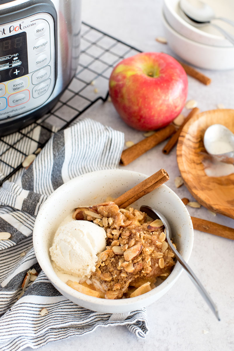 a 45 degree shot of Instant Pot apple crisp in a white bowl, with a scoop of vanilla ice cream on top, with an Instant Pot, an apple, and cinnamon sticks in the background