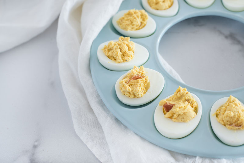 Blue circular egg tray filled with hard-boiled eggs made in the Instant Pot / pressure cooker deviled eggs with BBQ sauce and bacon crumbles.