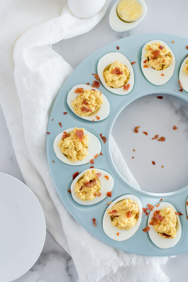 Overhead of a blue round egg tray with Instant Pot BBQ Deviled eggs with crumbled crispy bacon on a white marble background with a white dishcloth and a white plate.
