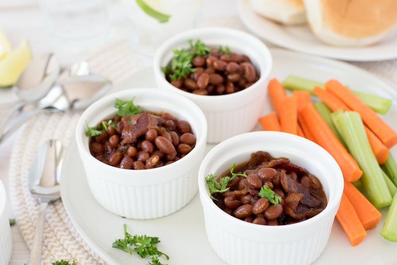 Three white ramekins filled with Instant Pot baked beans served with celery and carrot sticks on the side