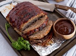 BBQ bacon Instant Pot meatloaf, sliced and ready to serve, pictured on a broiling pan with garnish, a knife, and extra BBQ sauce.