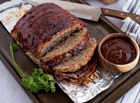 BBQ bacon Instant Pot meatloaf, sliced and ready to serve, pictured on a broiling pan with garnish, a knife, and extra BBQ sauce.