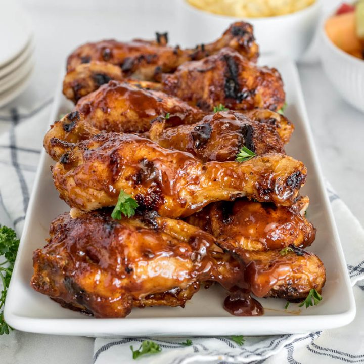 A head-on image of a rectangular white platter of BBQ chicken legs, with a fruit salad in the background