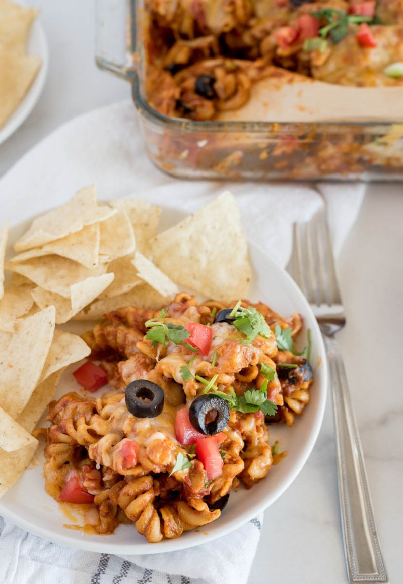 Instant Pot chicken enchilada pasta topped with sliced olives, diced tomatoes, and fresh cilantro on a plate with chips, placed in front of a serving dish.
