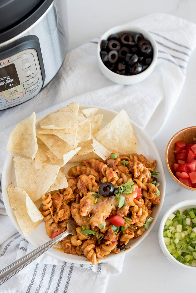 Pressure Cooker chicken enchilada pasta served on a plate with chips, with diced green onions, tomatoes, and sliced olives in bowls in front of an Instant Pot.