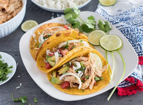 A plate of three Instant Pot chicken tacos, filled with rice, cilantro, avocado, and diced tomato, garnished with two lime slices and a spring of cilantro