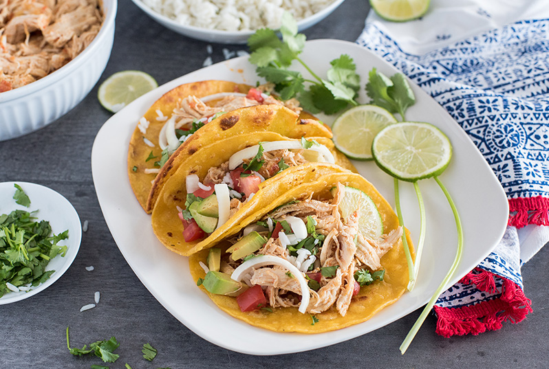 A plate of three Instant Pot chicken tacos, filled with rice, cilantro, avocado, and diced tomato, garnished with two lime slices and a spring of cilantro