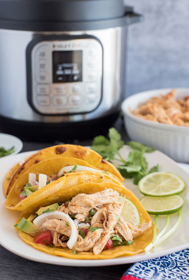 Three Pressure Cooker Chicken Tacos plated with a garnish of cilantro and limes, with an Instant Pot and a bowl of shredded chicken taco meat in the background