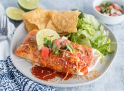 White dinner plate filled with tortilla chips, romaine lettuce and an Instant Pot Chili Colorado Smothered Burrito topped with melty cheese, lime wedge, fresh salsa and more enchilada sauce on a white background with a blue and white napkin and a silver fork.