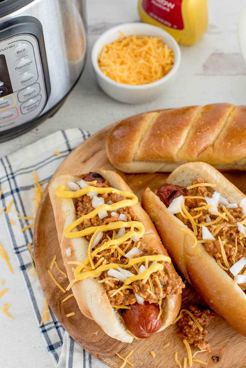 Instant Pot Chili Dog sauce, served on two different hot dogs, garnished with mustard, cheese, and onions, and shown on a wooden platter, with an INstant Pot in the background