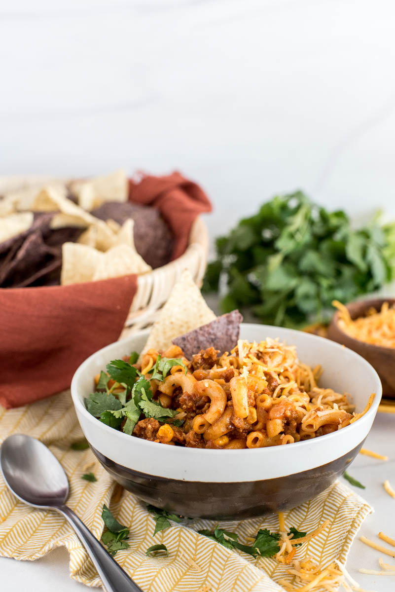 A black and white bowl of Instant Pot chili mac, garnished with cilantro and blue and white corn chips, sitting on a yellow napkin with a spoon, with a basket of chips, additional cilantro, and cheese visible in the background