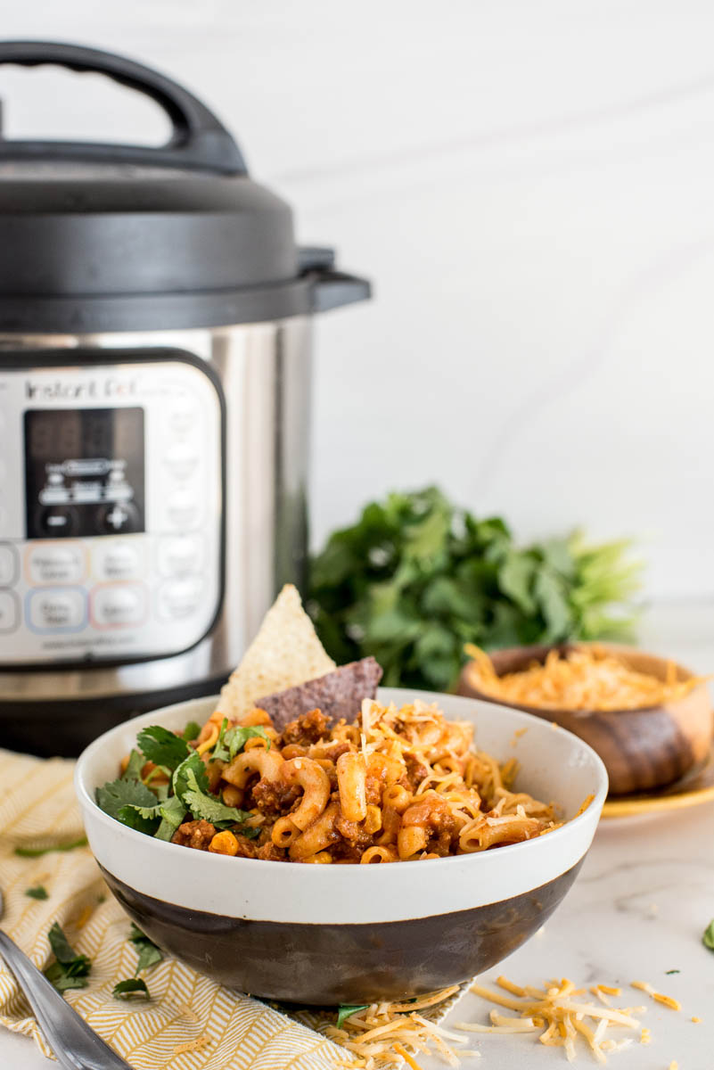 a direct shot of a black and white bowl of Instant Pot Chili Pasta with a cilantro garnish and white and blue corn chips, with an Instant Pot visible in the background