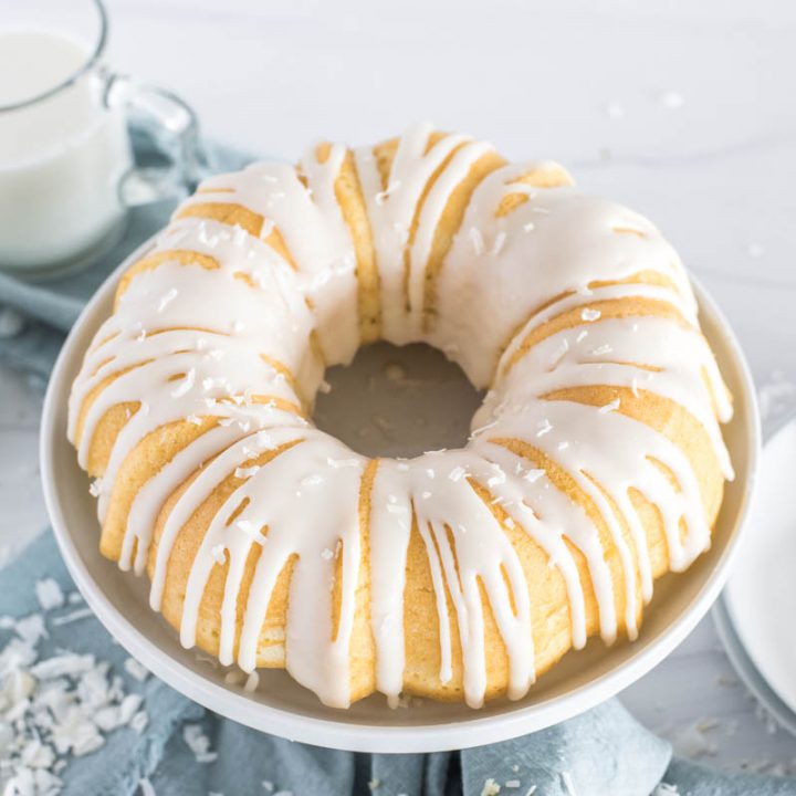 Close-up 45 degree shot of the Instant Pot coconut bundt cake, drizzled in bright white icing, with a glass of milk, white plates and forks, and a blue napkin in the background