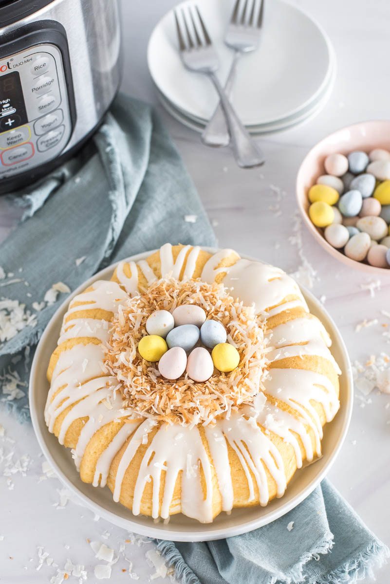 An overhead shot of the Instant Pot coconut bundt cake dressed up for Easter, with a toasted coconut "birds nest" in the middle center of the bundt, filled with pasted Cadbury Mini Eggs, with a blue napkin, white plates and forks, an Instant Pot, and a pastel pink bowl of Cadbury Mini Eggs visible in the background