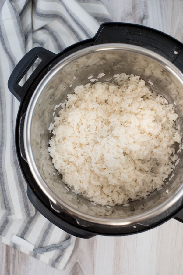 Overhead of an electric pressure cooker filled with fluffy white coconut rice on a light wooden background with a grey and white striped linen