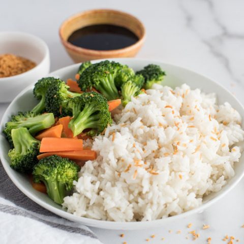 Large wooden dish filled with instant Pot coconut rice topped with toasted coconut flakes and served with steamed green broccoli florets, orange carrot sticks on a white and grey marble background next to a pair of wooden chopsticks