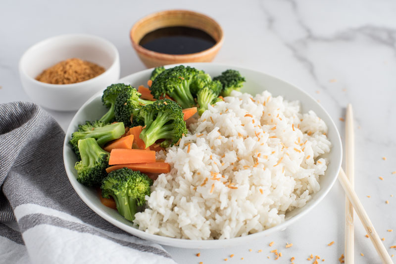 Large wooden dish filled with instant Pot coconut rice topped with toasted coconut flakes and served with steamed green broccoli florets, orange carrot sticks on a white and grey marble background next to a pair of wooden chopsticks