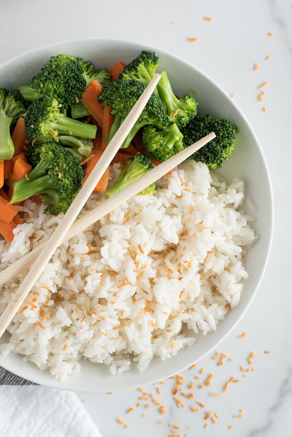 close up on a white dish of instant pot coconut rice topped with toasted coconut flakes with steamed broccoli and carrots and two wooden chopsticks crossed on top of the dish