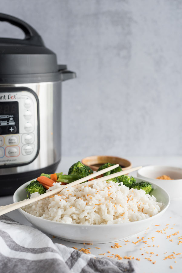 Side view of a white bowl of instant pot coconut rice topped with toasted coconut flakes next to steamed broccoli and carrots on a white and grey background in front of an instant pot pressure cooker