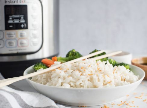 Side view of a white dish filled with instant Pot coconut rice topped with toasted coconut flakes and wooden chopsticks in front of an electric pressure cooker.