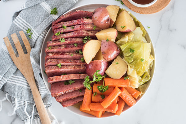 Corned Beef and Cabbage on a white circular serving dish with a wooden spoon resting on a grey and white linen napkin.
