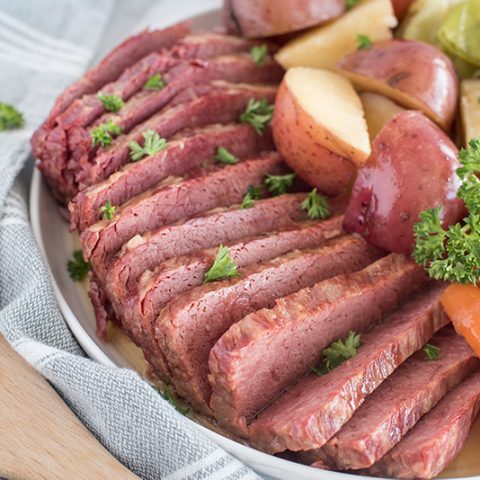45 degree shot of thin slices of Instant Pot corned beef served with tender carrots, white potatoes, and cabbage, garnished with fresh parsley on a round white serving dish