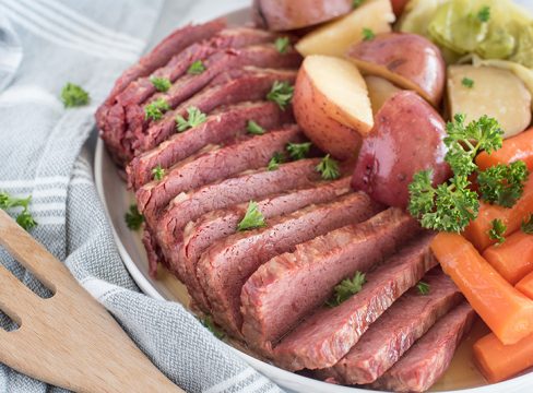 45 degree shot of thin slices of Instant Pot corned beef served with tender carrots, white potatoes, and cabbage, garnished with fresh parsley on a round white serving dish